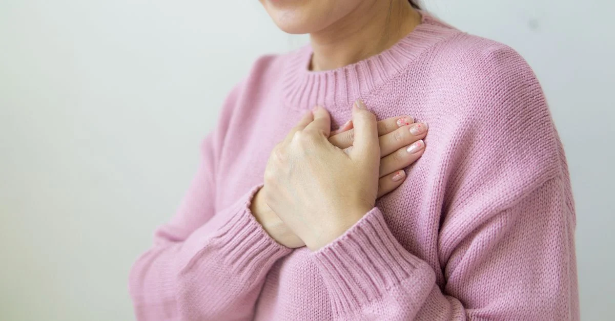 Une femme sereine en pull rose tient son cœur, symbolisant la chaleur et le soin.
