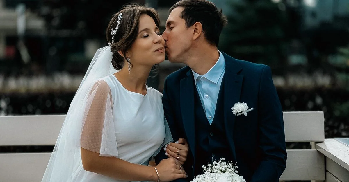 Les mariés partagent un tendre baiser sur un banc de parc lors d'une séance photo le jour du mariage, créant un moment romantique.
