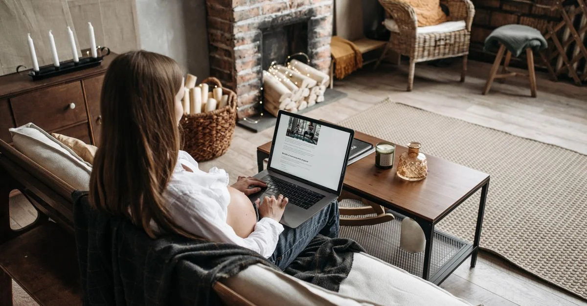 Femme enceinte travaillant à distance depuis chez elle, assise sur un canapé avec un ordinateur portable, intérieur chaleureux.