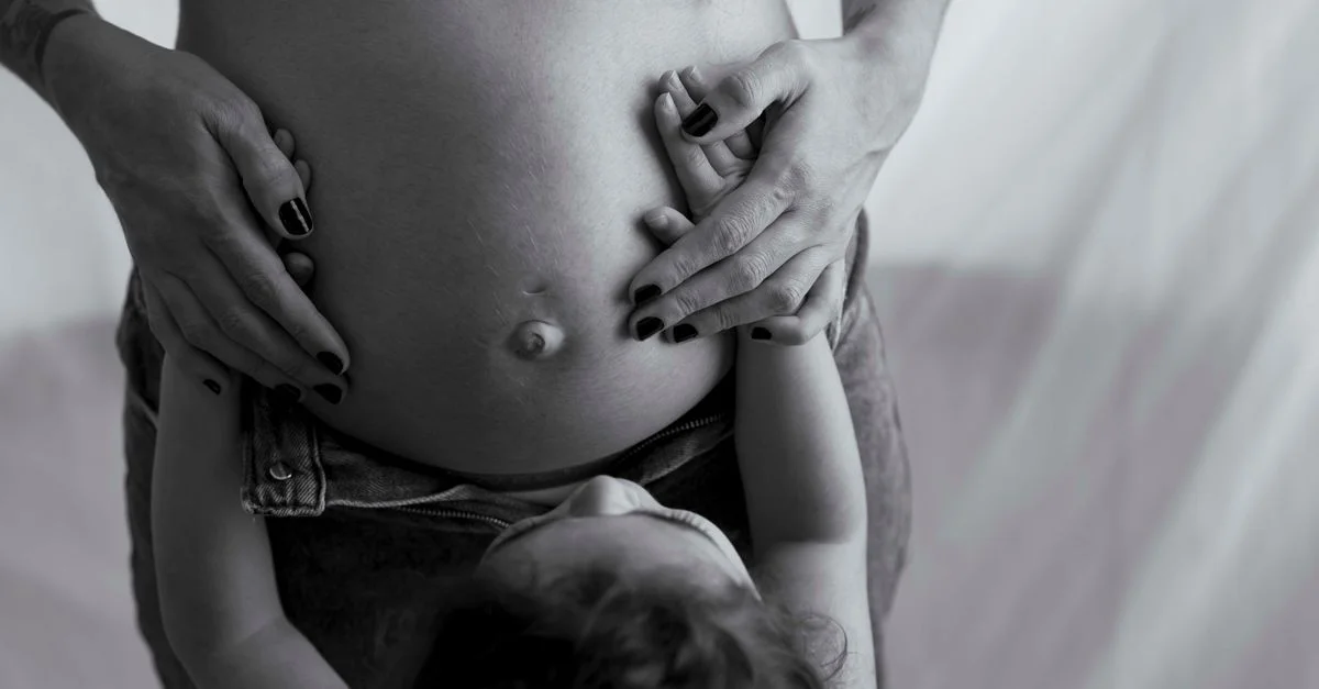 Photo en noir et blanc capturant les mains d'un enfant sur un ventre de femme enceinte, symbolisant le lien familial.