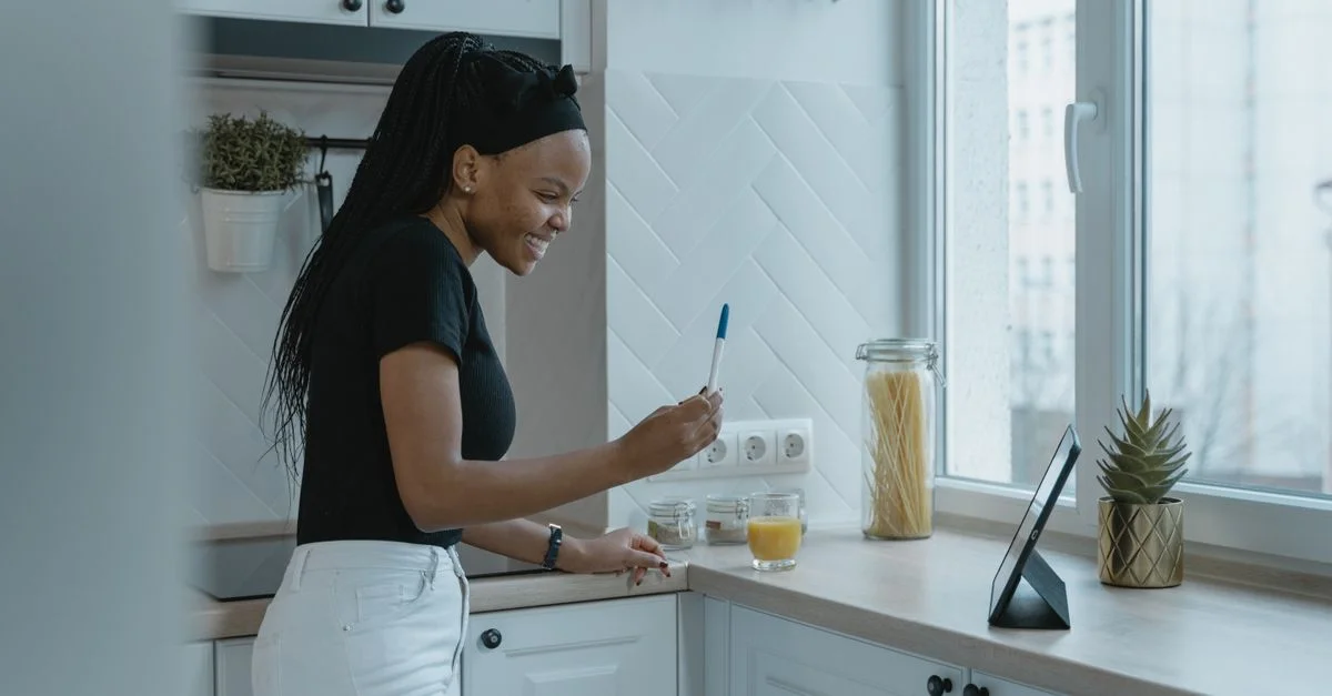 Une femme heureuse souriant tout en tenant un test de grossesse dans sa cuisine, exprimant joie et anticipation.