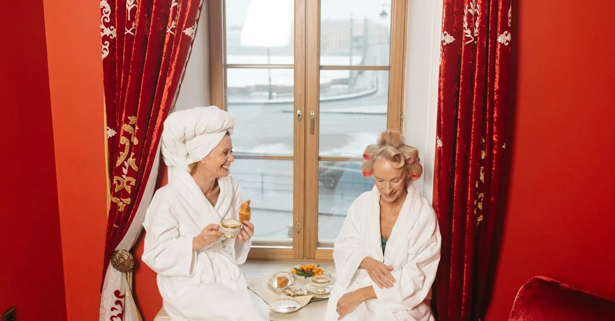 Deux femmes âgées en peignoir profitent d'un petit déjeuner près d'une fenêtre dans une chambre d'hôtel cosy.