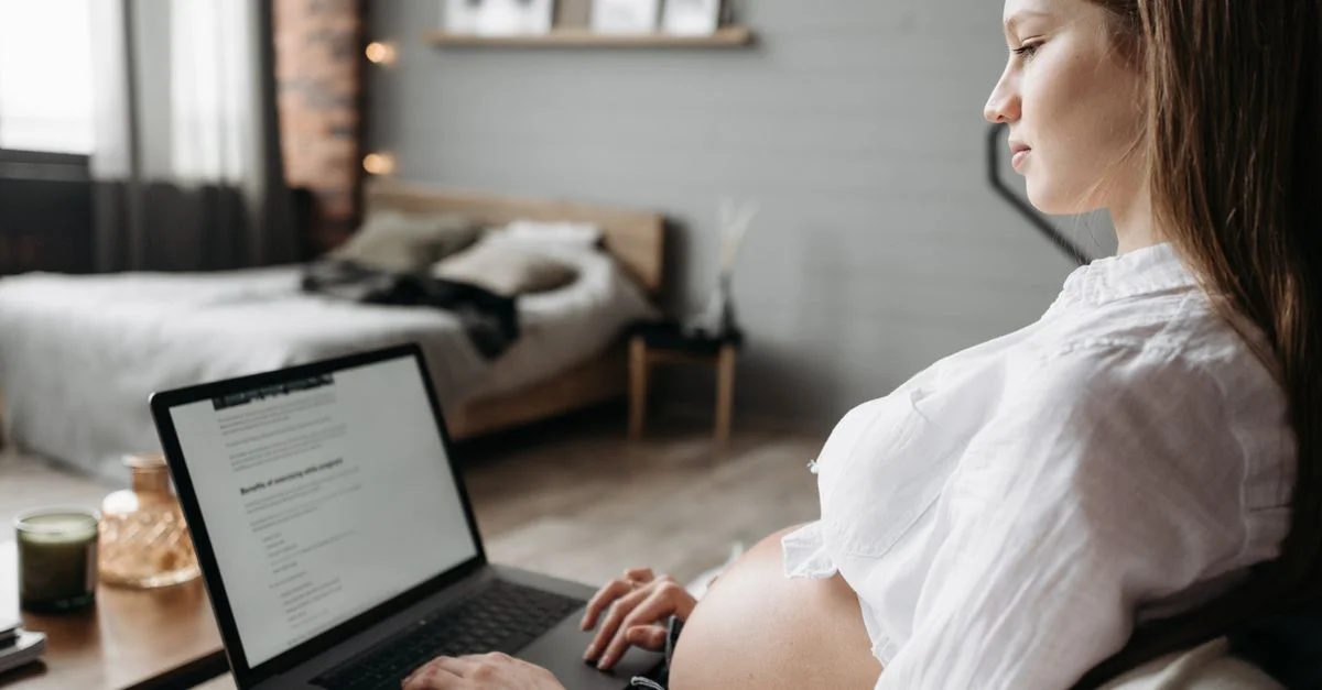 Femme enceinte travaillant sur un ordinateur portable dans une chambre confortable et lumineuse. Vue latérale de son ventre arrondi.