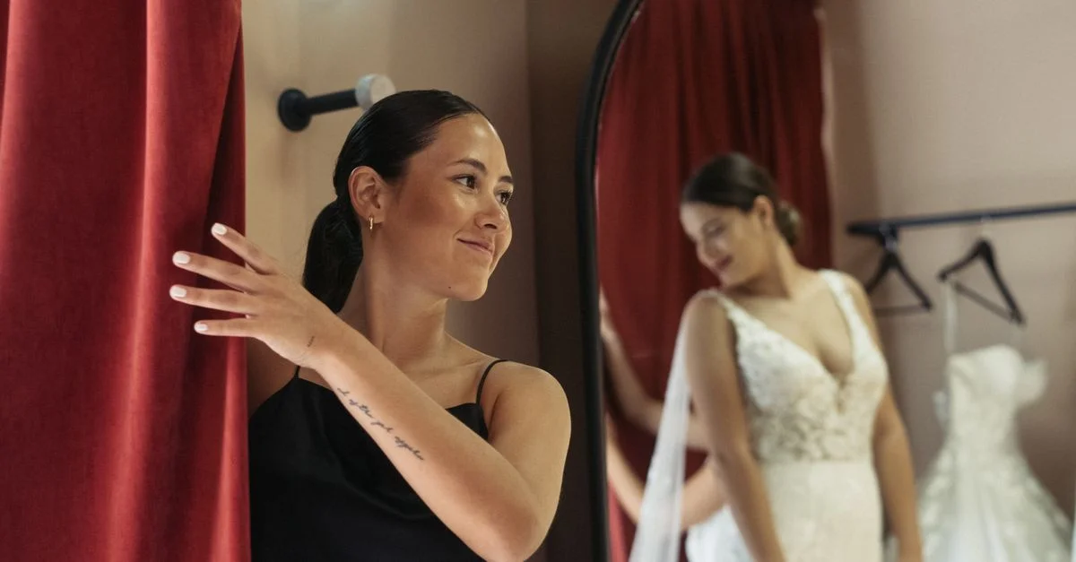 Femme dans une cabine d'essayage avec une robe de mariée, souriant devant un miroir avec un rideau rouge.