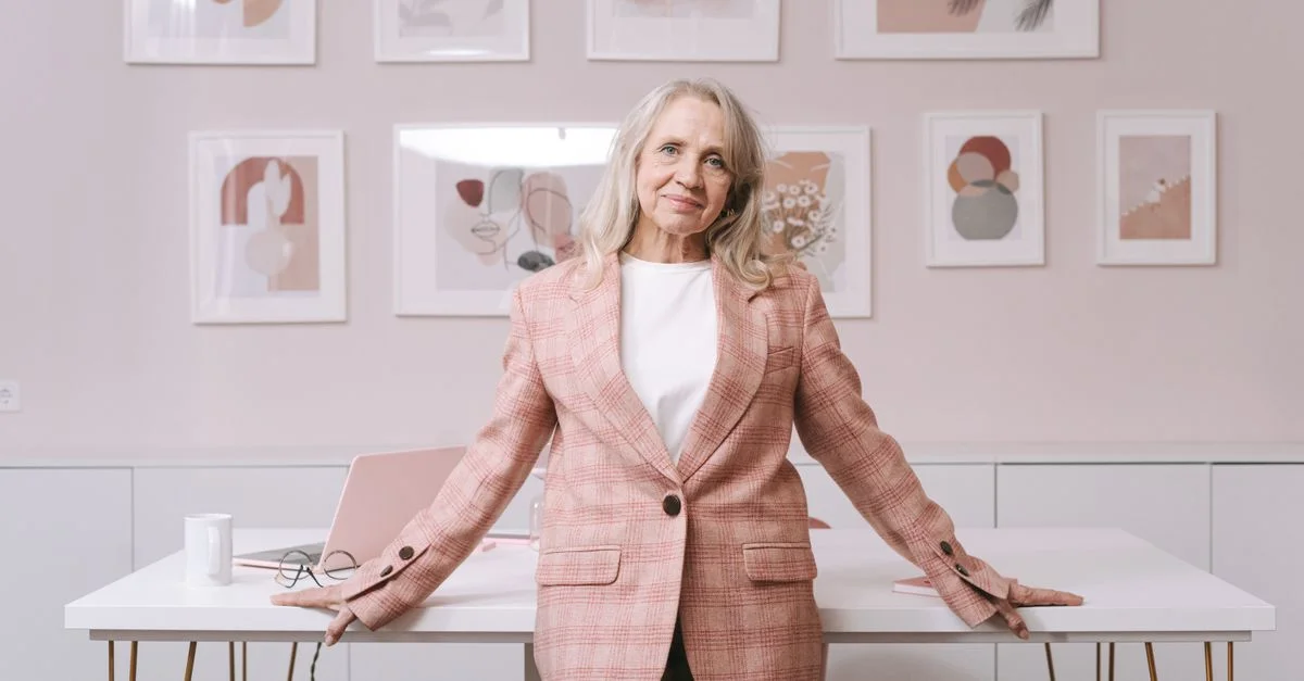 Femme âgée en blazer rose posant avec confiance dans un environnement de bureau à domicile élégant.