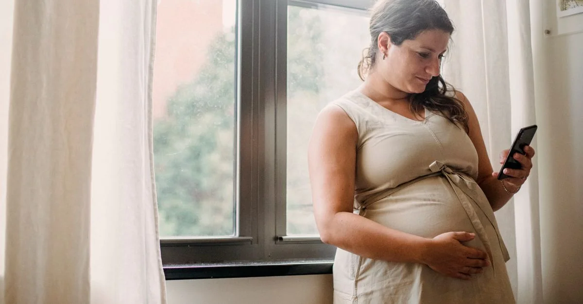 Femme enceinte se tenant près de la fenêtre, naviguant sur son smartphone dans un cadre domestique calme.